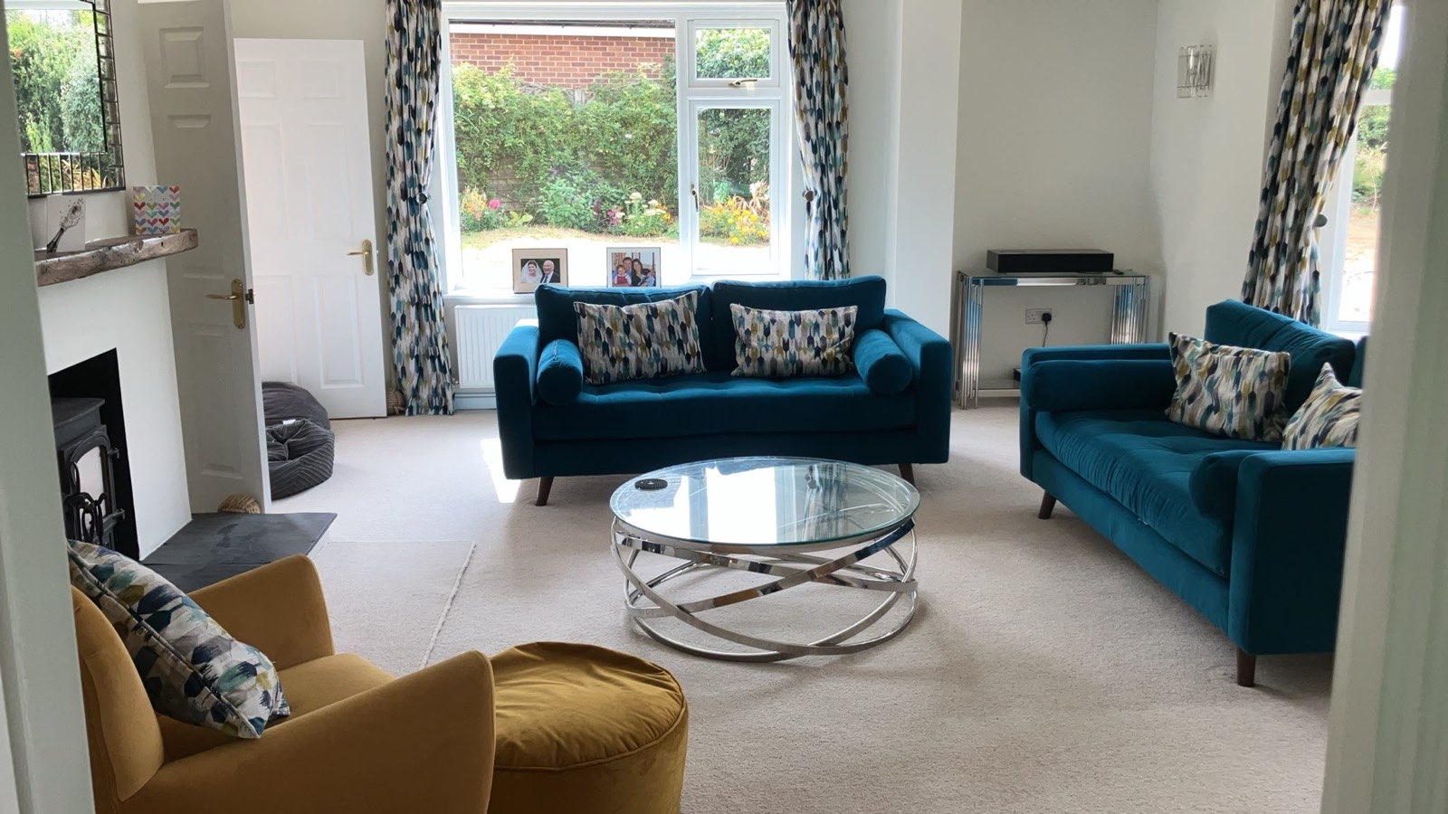 View into the living room from the hallway showing the wood burner, both teal sofas, the ochre armchair and footstool, and the patterned curtains