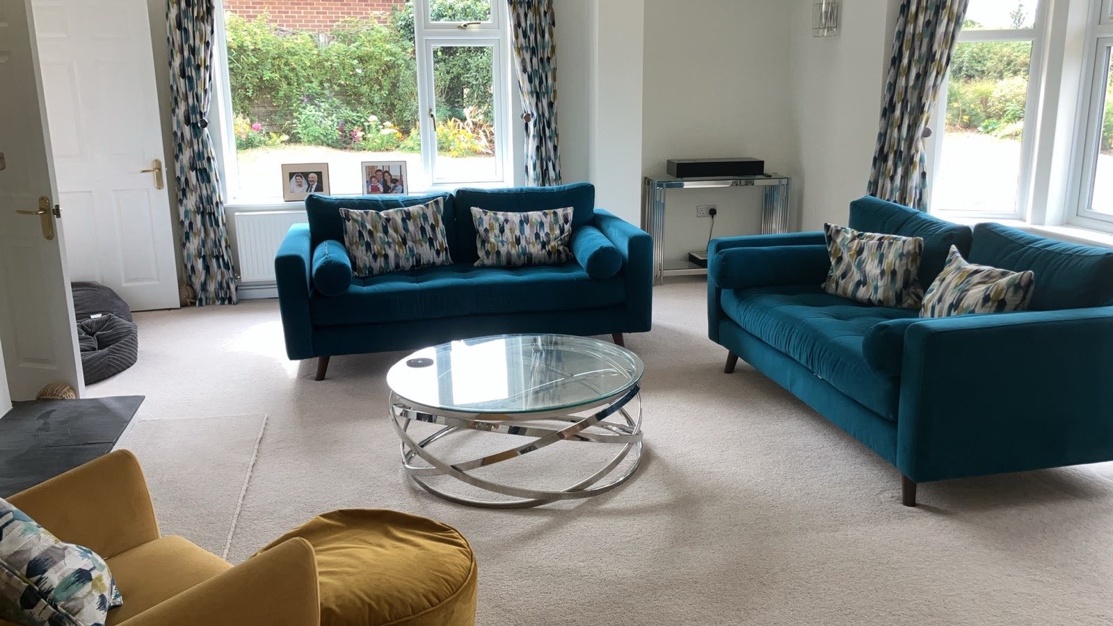 Living room with two teal velvet sofas facing each other across a round glass and chrome coffee table, patterned curtains at the windows and an ochre armchair in the foreground