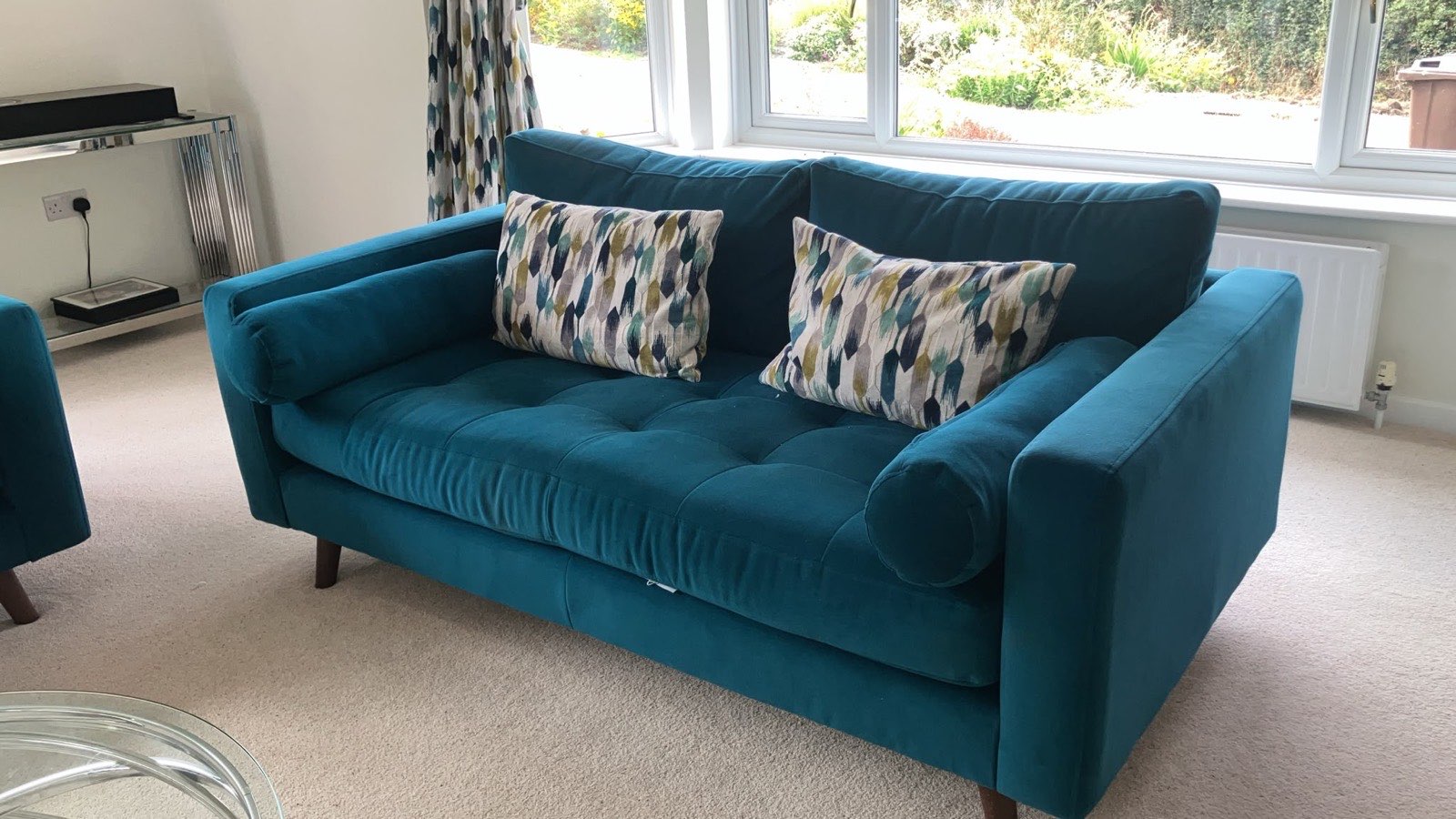 Close view of a teal velvet sofa with two patterned cut velvet cushions, next to the matching patterned curtain